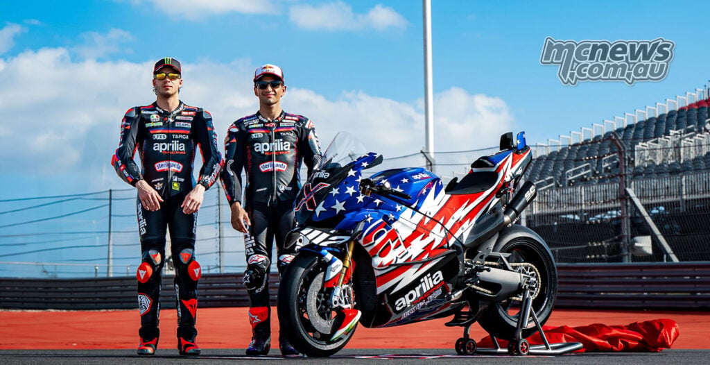 Two Aprilia Racing riders stand beside the Aprilia X 250TH under a large American flag at Circuit of the Americas.