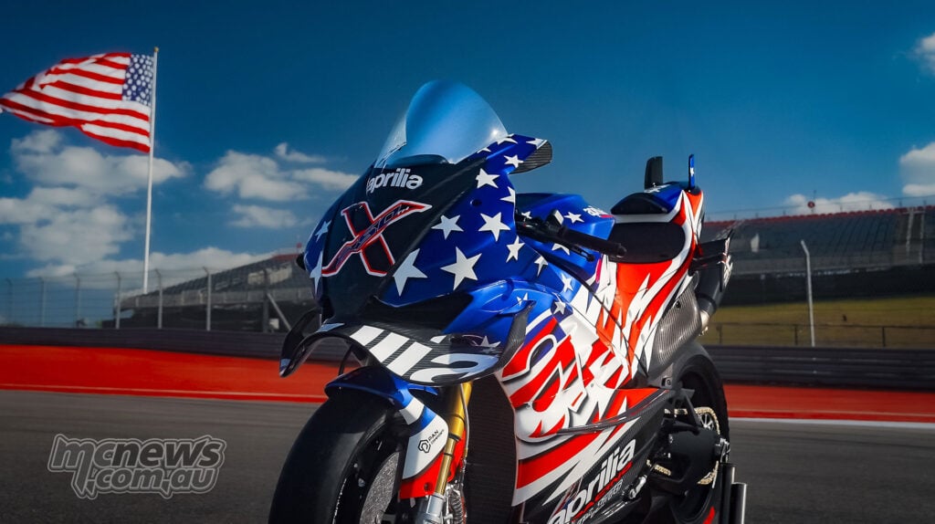 Front three-quarter image of the Aprilia X 250TH at COTA with a blurred American flag and grandstand behind it.