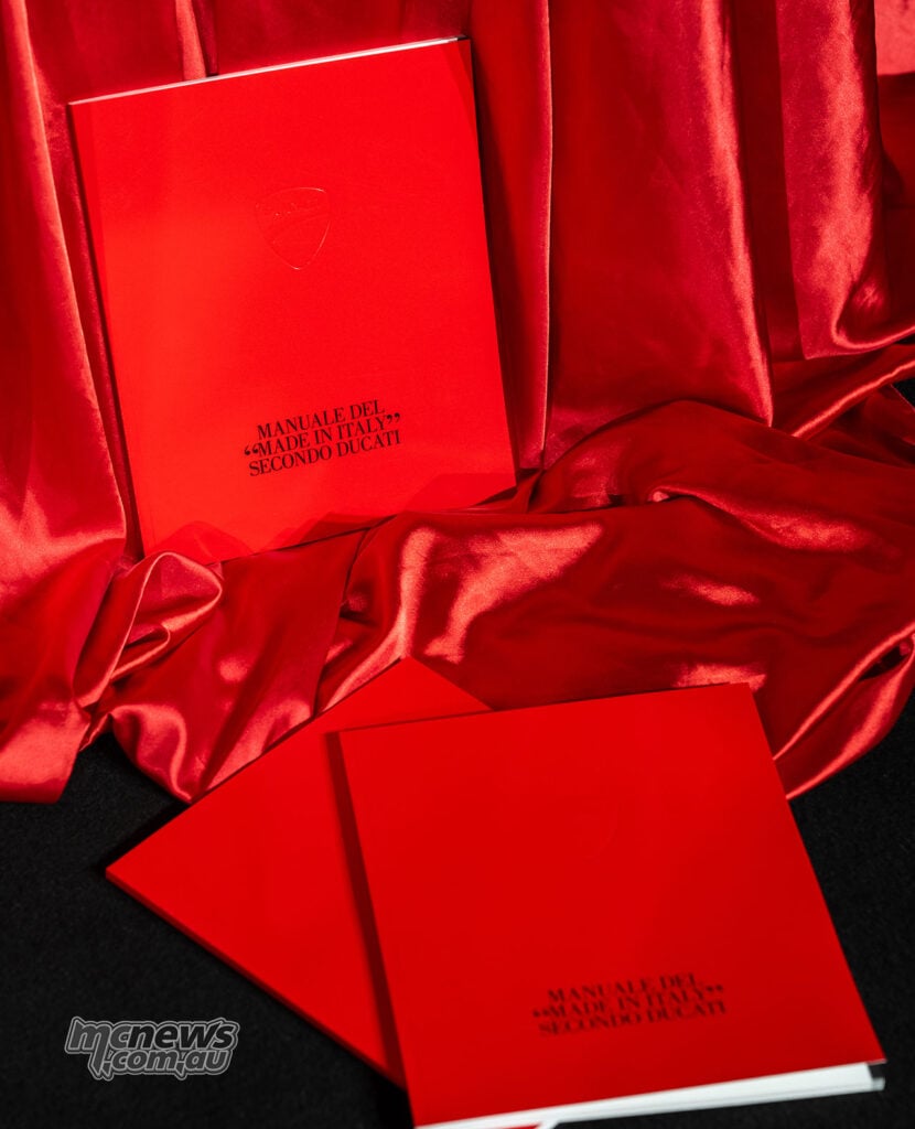 Red Ducati centenary handbook displayed on a red satin backdrop.
