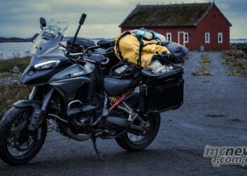 Ducati Multistrada V4 packed with touring luggage on a coastal road near a red building in northern Europe.
