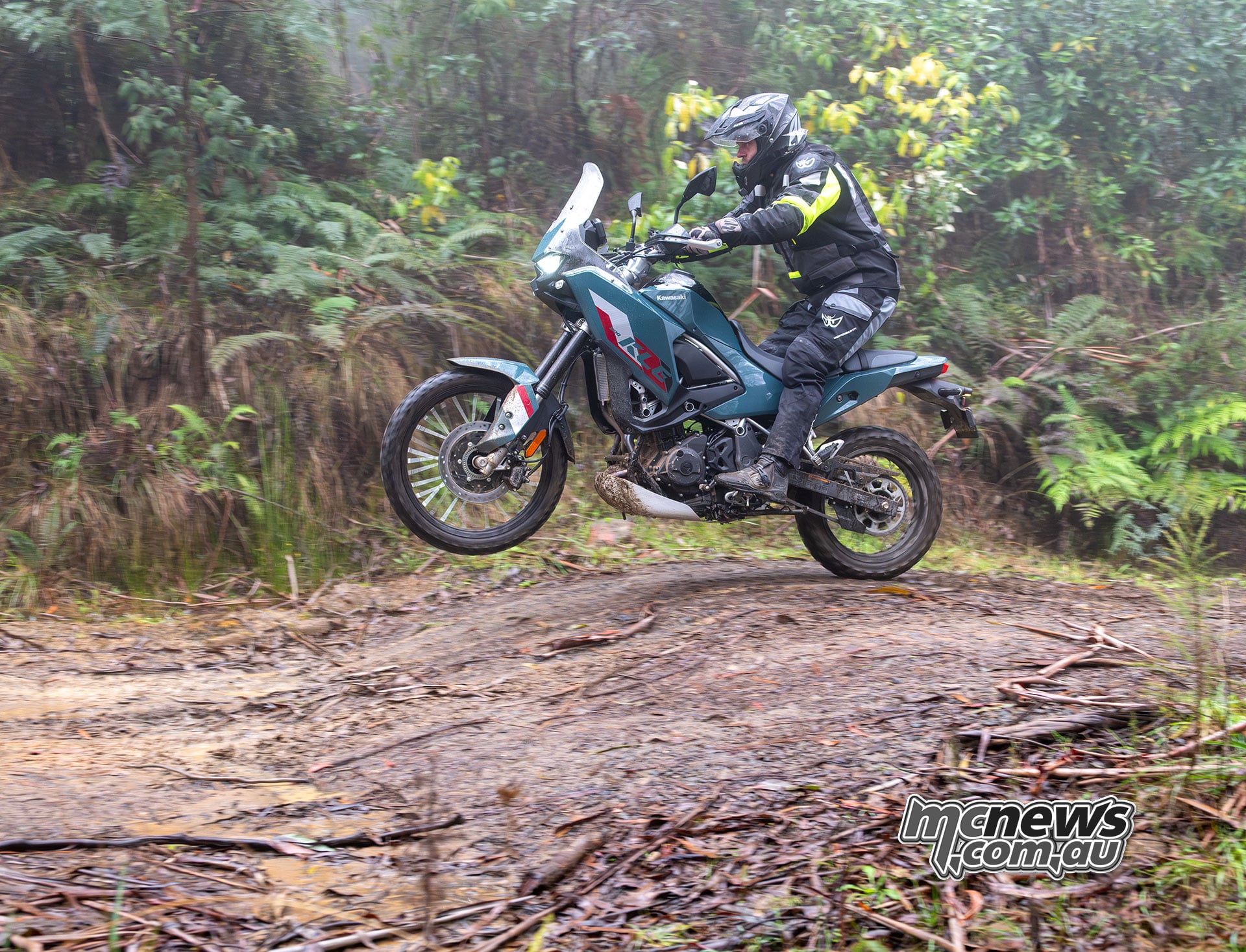 Rider standing on the footpegs of a Kawasaki KLE500 SE with both wheels airborne above a muddy forest trail.