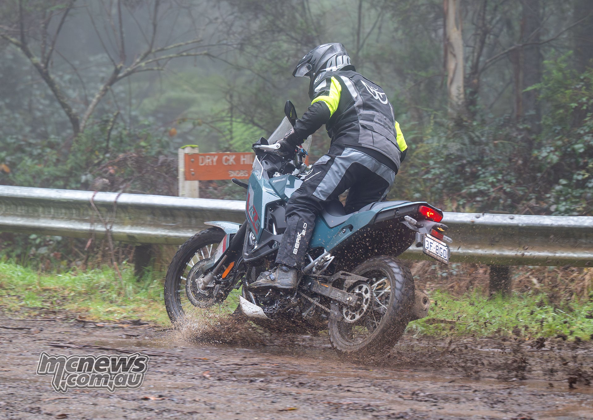 Rear three-quarter view of a rider turning a Kawasaki KLE500 SE on a muddy track, with dirt spraying from the rear tyre.