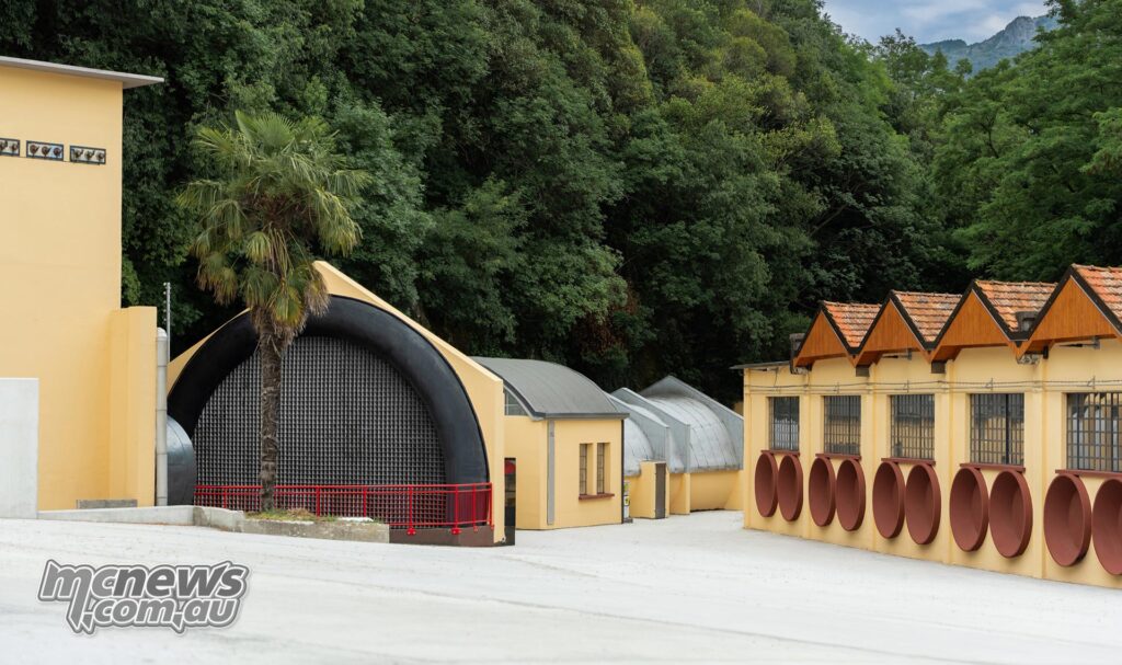 The historic yellow Moto Guzzi factory buildings in Mandello del Lario, surrounded by trees and mountains.