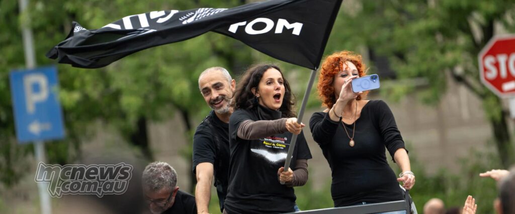 Three people attending a Moto Guzzi event wave a black flag and film the crowd from a high platform.