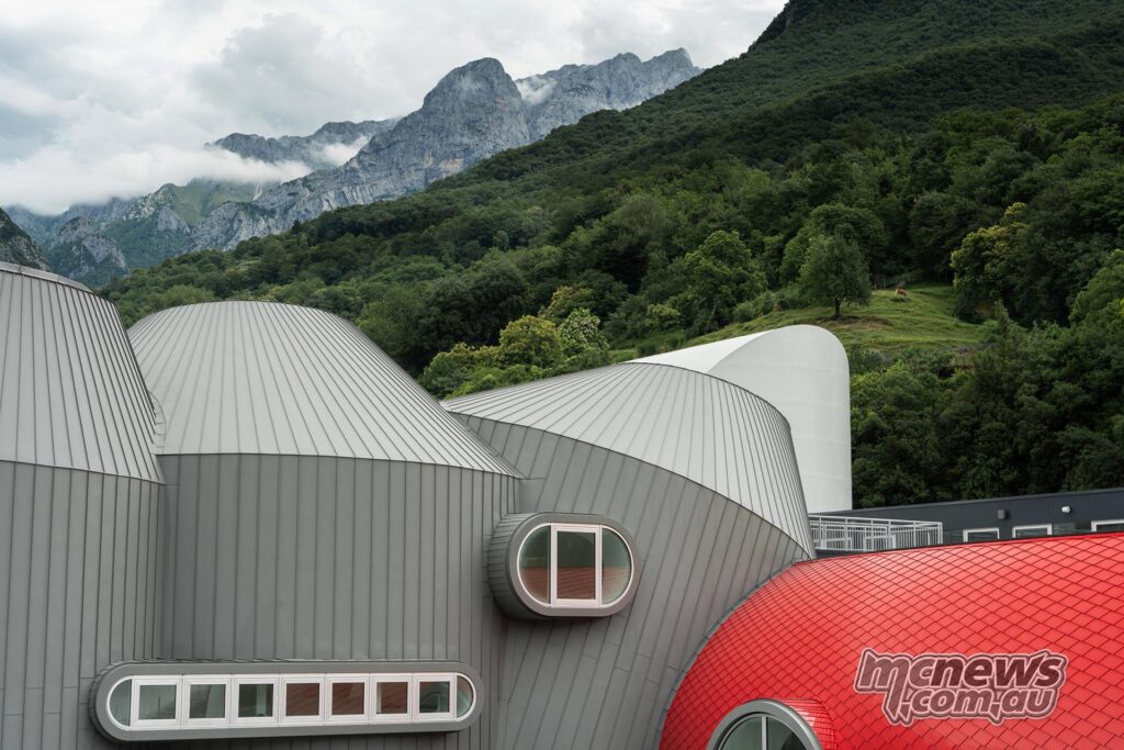 The winding brown and red buildings of the redeveloped Moto Guzzi campus and the wooded mountains behind them.