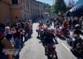 A large procession of Moto Guzzi motorcycles and sidecars riding between crowds lining a street in Mandello del Lario.
