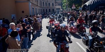 A large procession of Moto Guzzi motorcycles and sidecars riding between crowds lining a street in Mandello del Lario.