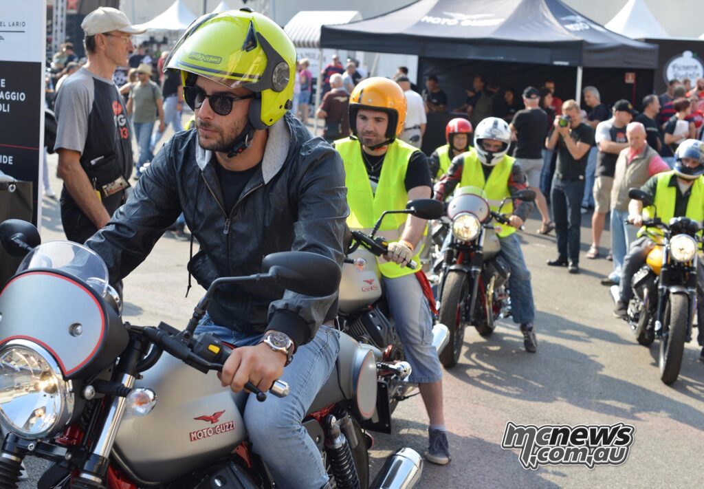 Moto Guzzi riders wearing helmets and high-visibility jackets line up for a test ride at a busy motorcycle event.