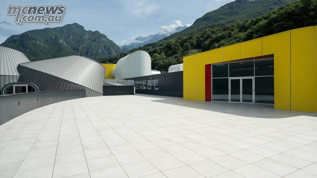 Open courtyard at the redeveloped Moto Guzzi site with yellow and brown buildings set beneath forested mountains.