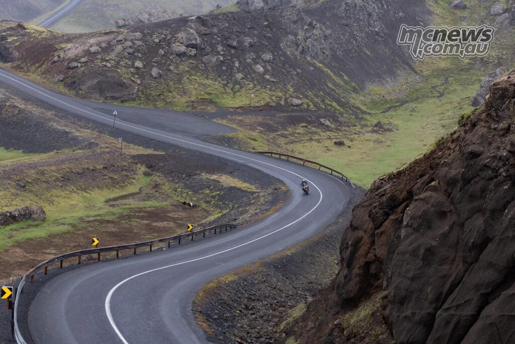 Motorcyclist riding a Norton Atlas around a sweeping road through rocky Icelandic hills.