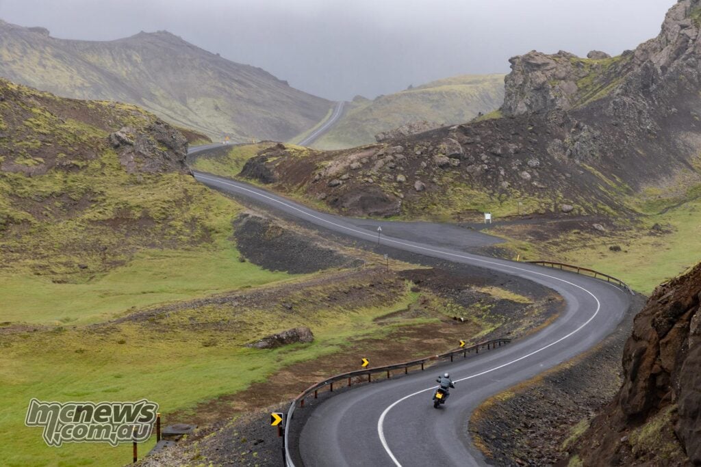 Motorcyclist riding a Norton Atlas along a winding road through Icelandic volcanic hills.