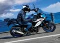 2027 Suzuki SV-7GX in blue and white being ridden along a coastal road, showing its crossover bodywork, windscreen, rear carrier and exposed V-twin engine.