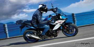 2027 Suzuki SV-7GX in blue and white being ridden along a coastal road, showing its crossover bodywork, windscreen, rear carrier and exposed V-twin engine.