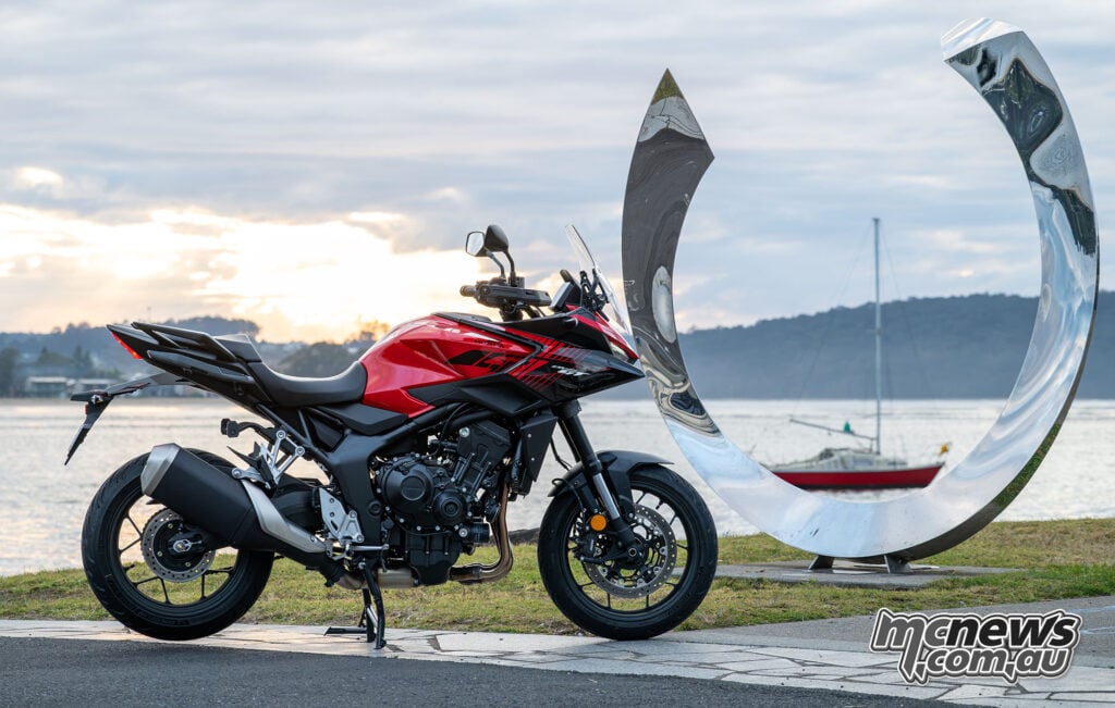 Red Honda CB1000GT parked beside a polished waterfront sculpture in evening light.
