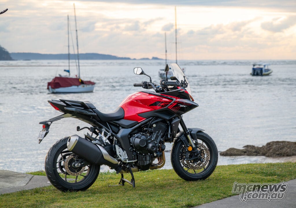 Red Honda CB1000GT without panniers parked beside the water with sailing boats behind it.