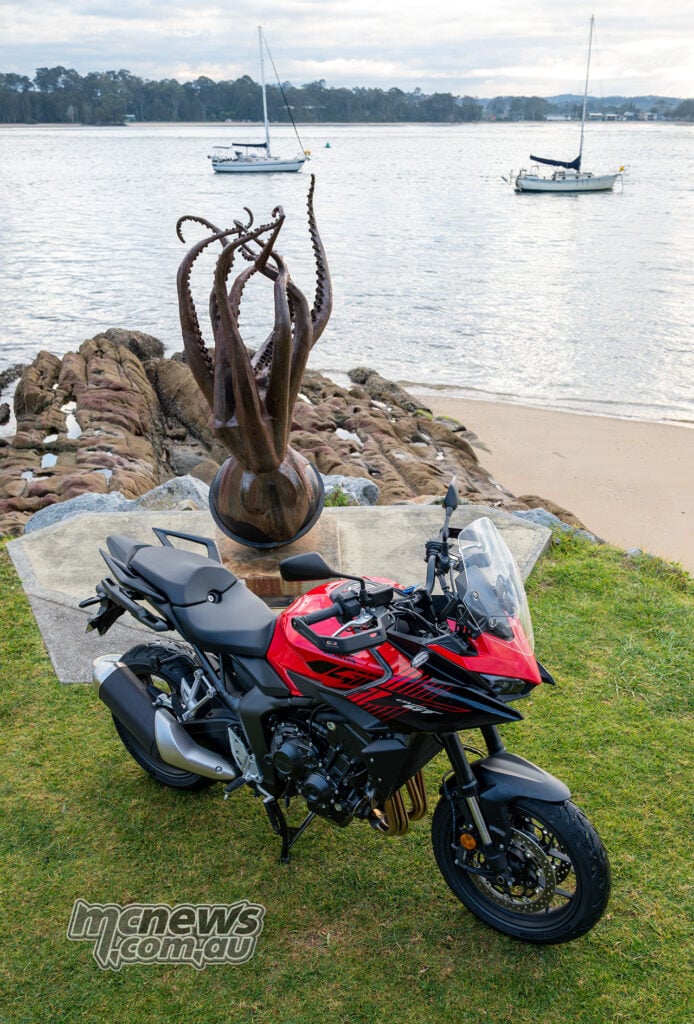 Honda CB1000GT parked below an octopus sculpture overlooking a harbour with yachts.