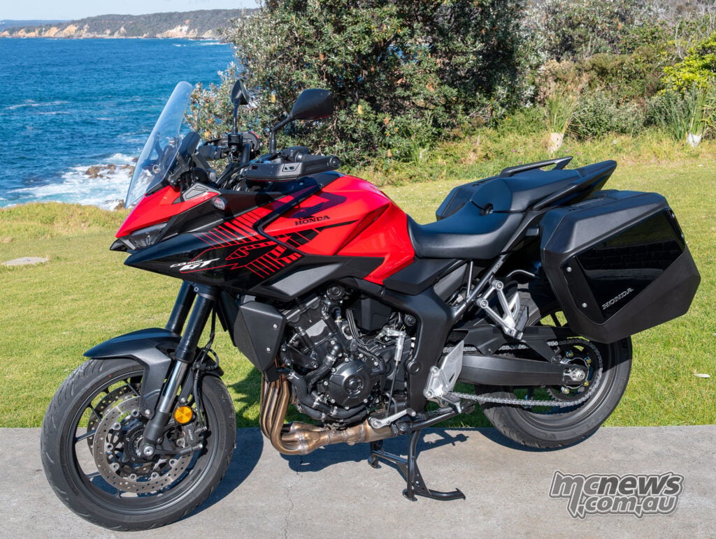Red Honda CB1000GT with panniers viewed from the front-left at a coastal lookout.