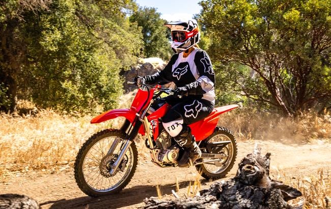 Rider in black and white riding gear on a red Honda CRF125F beside a fallen log on a dirt trail.