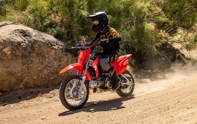 Young rider in black and yellow riding gear riding a Honda CRF110F past rocks on a dirt trail.