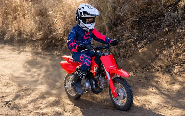 Young rider in blue and pink riding gear on a red Honda CRF50F along a dusty trail.