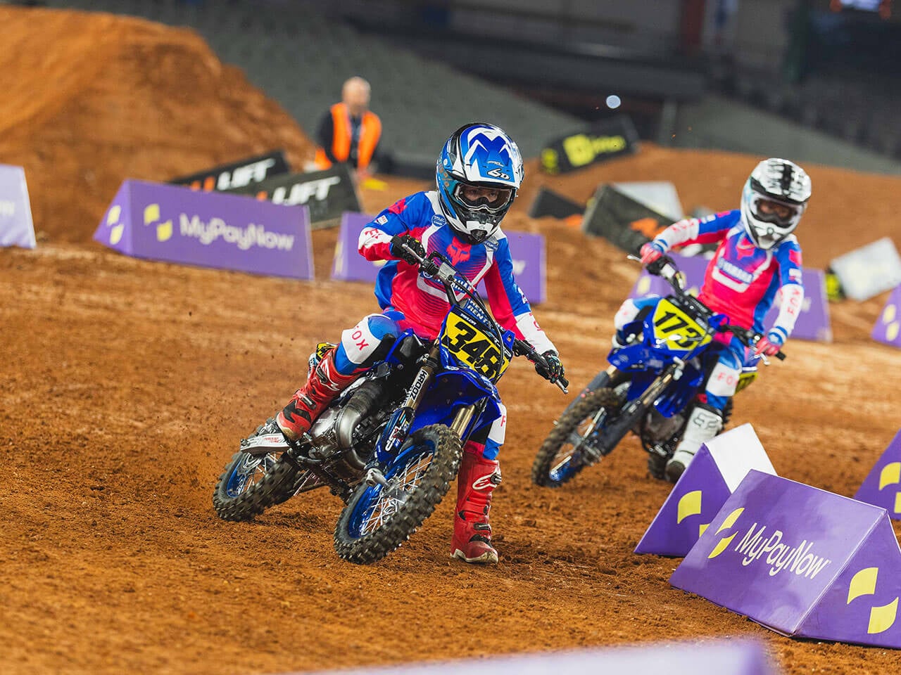 Two young Yamaha YZ65 riders racing through a turn on an indoor Supercross track