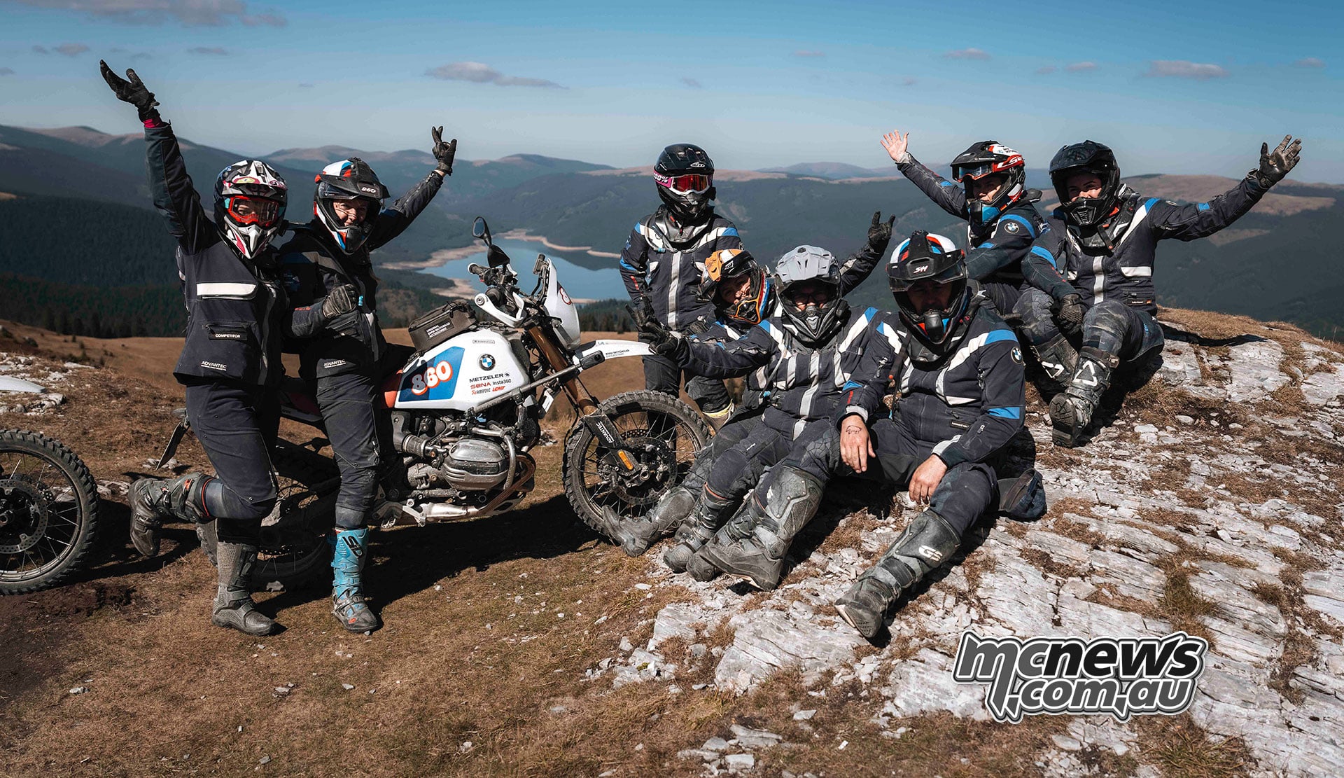 GS Trophy participants celebrate beside their BMW motorcycles on a rocky summit overlooking Romania’s mountains.