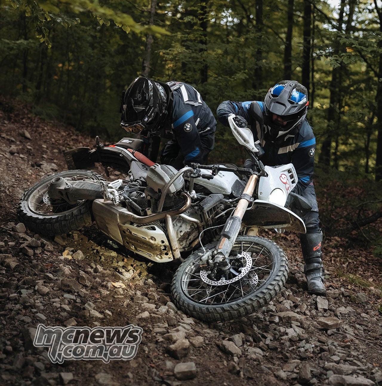 Two GS Trophy riders help recover a fallen BMW R 12 G/S on a steep, leaf-covered forest slope.