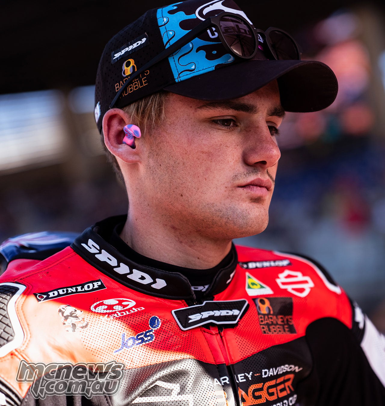 Close portrait of Archie McDonald in red and black leathers, with earplugs and sunglasses resting on his blue and pink cap.