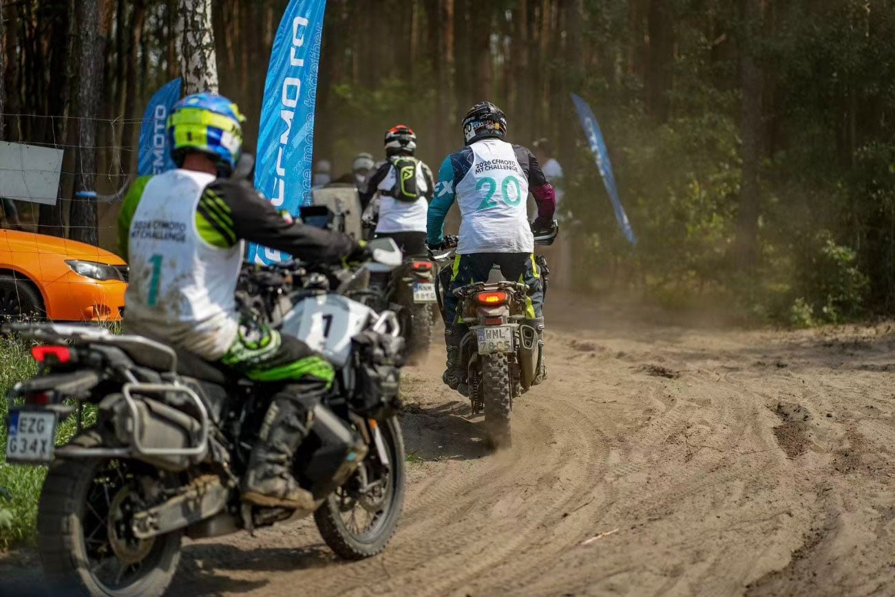 MT Challenge riders wearing numbered bibs follow a sandy track into woodland.