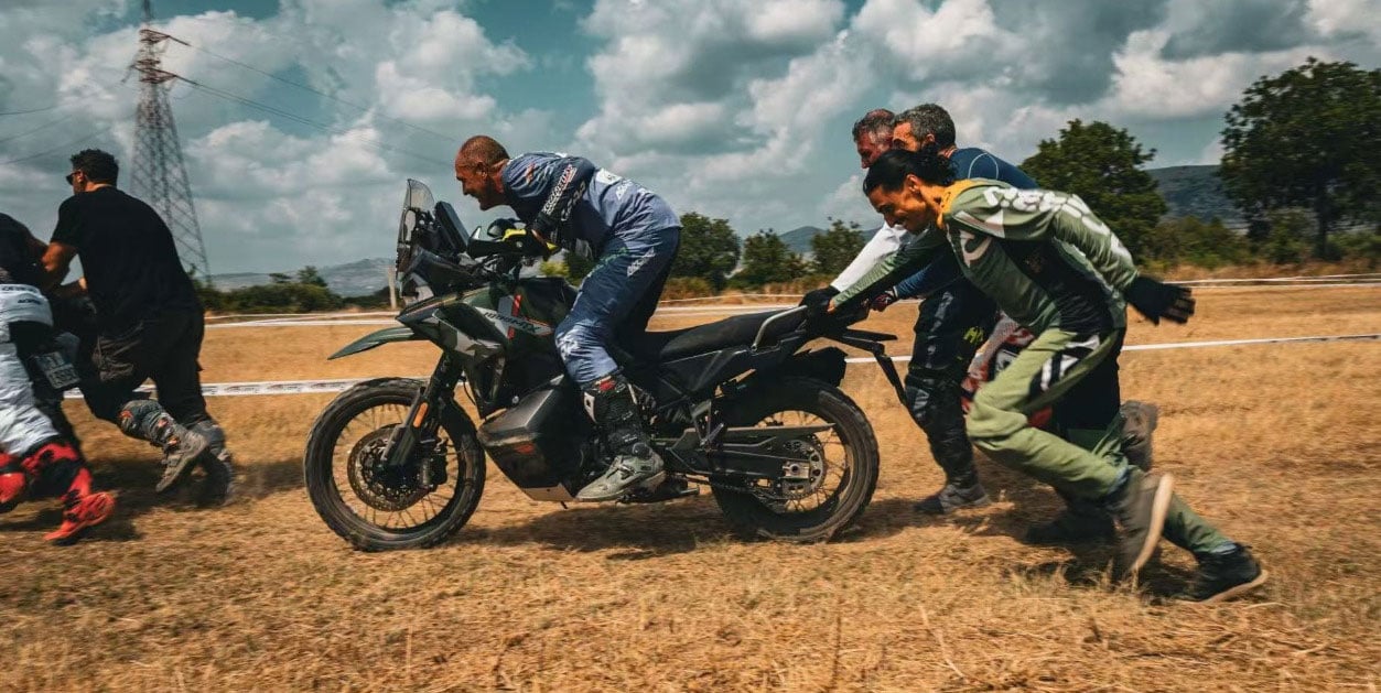 A group pushes an adventure motorcycle across a dry grass course with a rider aboard.