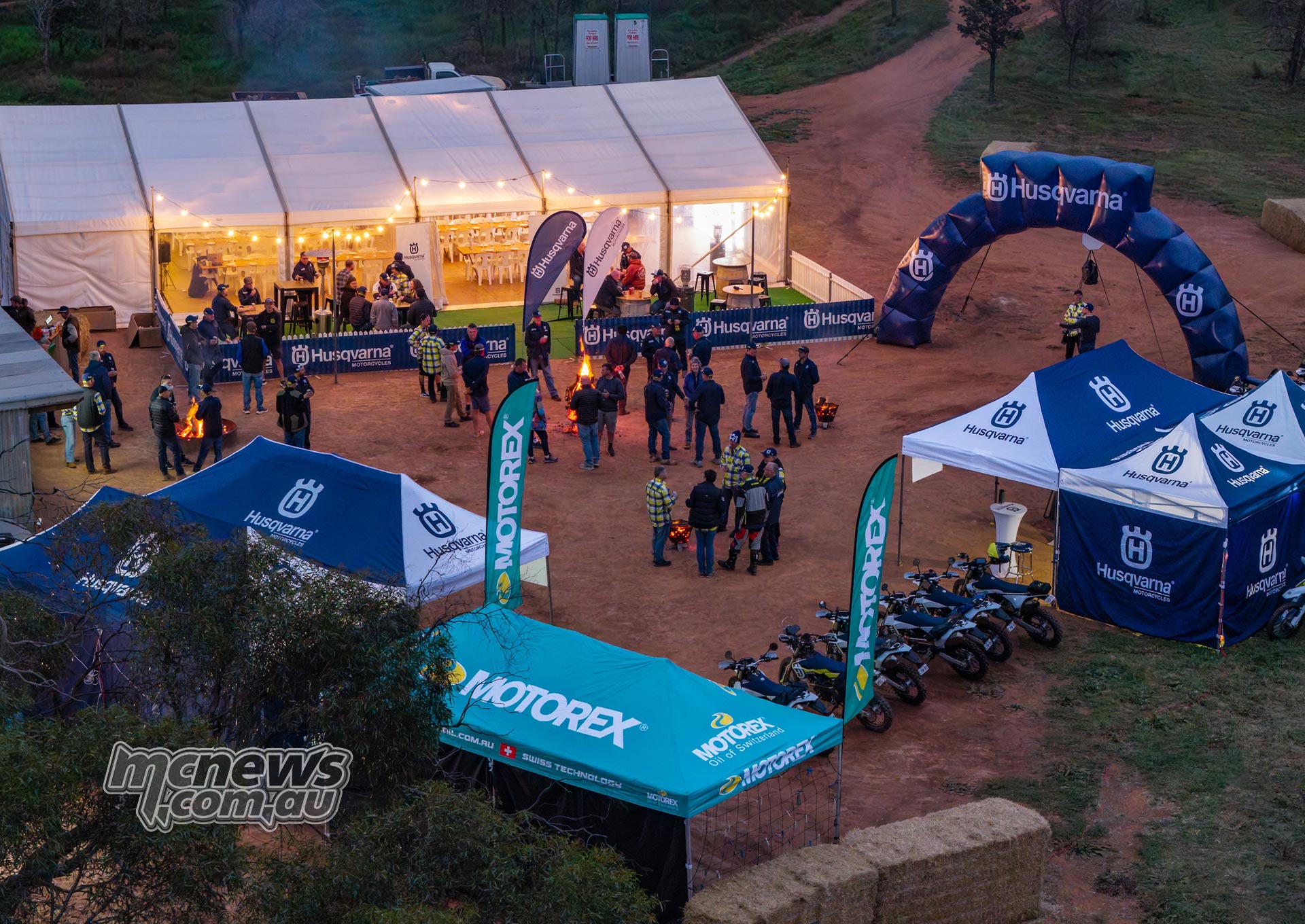 Riders gather beside an illuminated event marquee, Husqvarna tents and an inflatable arch at dusk.