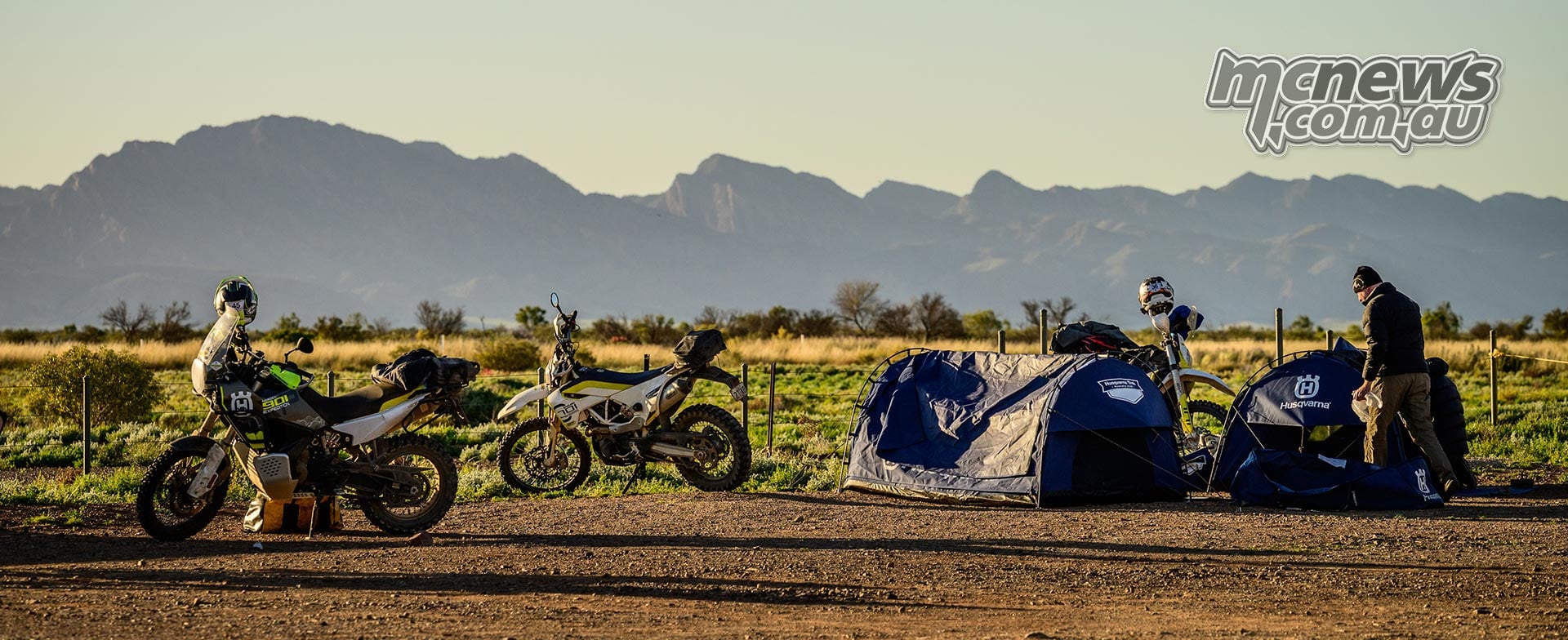 Husqvarna motorcycles parked beside blue swags, with a rider at camp and mountain silhouettes in the distance.
