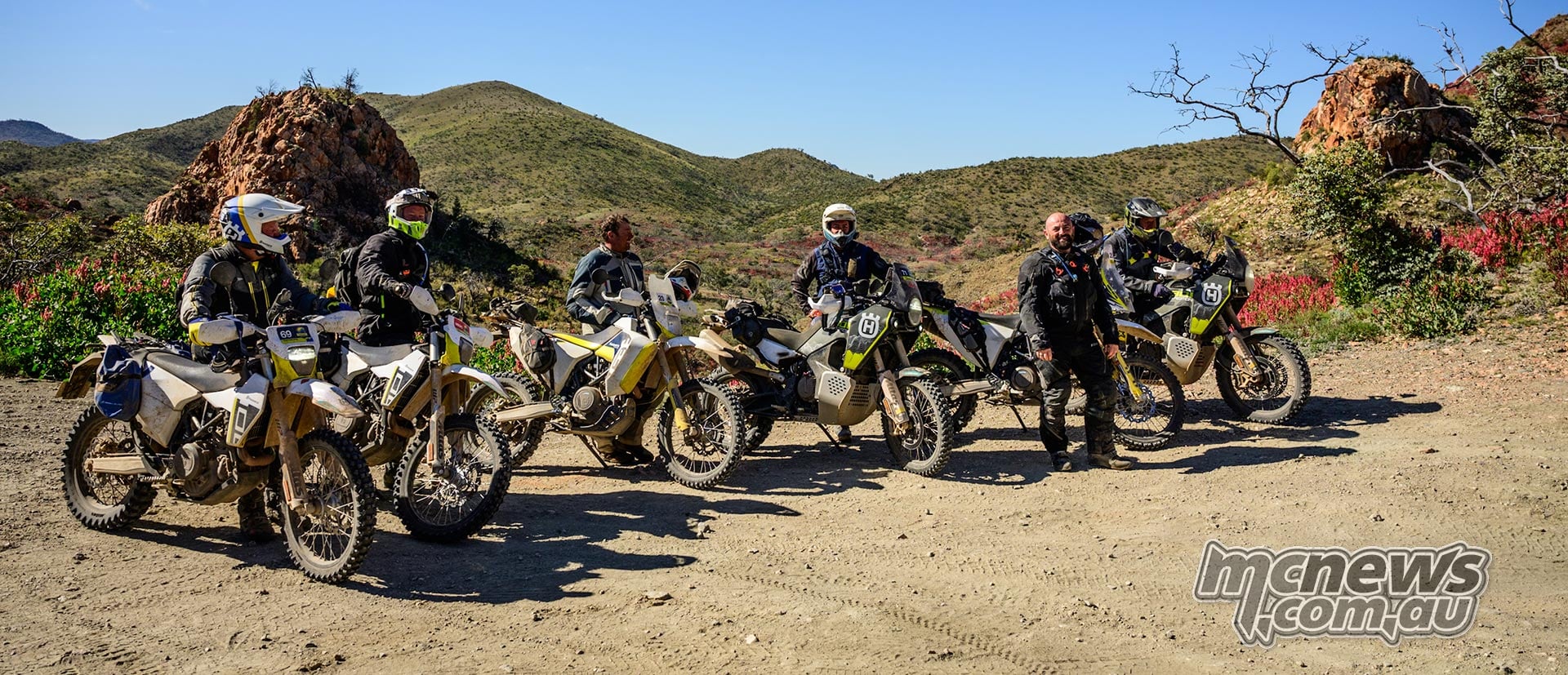 A group of Husqvarna riders poses with 701 Enduro and Norden motorcycles beside a rocky track.