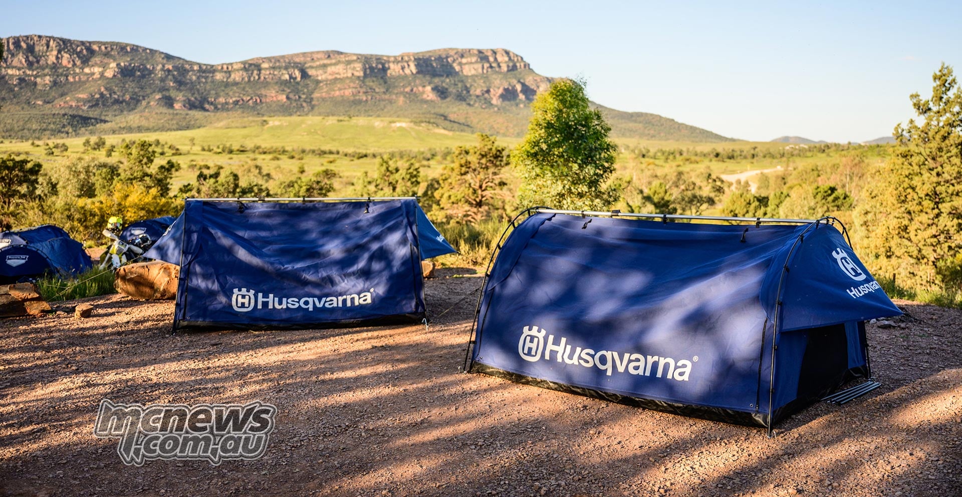 Blue Husqvarna swags pitched beneath trees, overlooking green country and a long rocky escarpment.