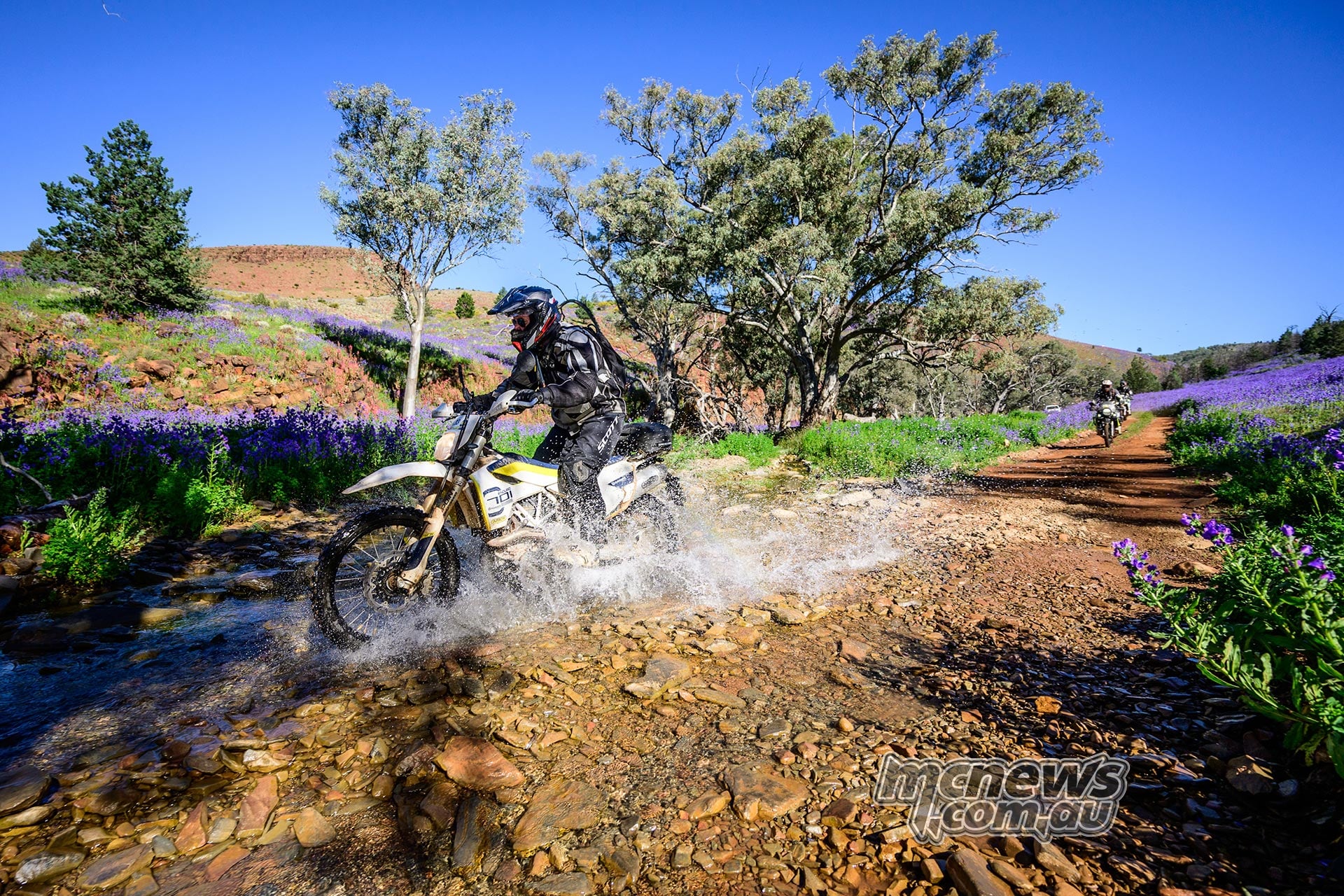 Husqvarna rider splashes through a shallow rocky creek beside purple wildflowers, with another rider approaching.