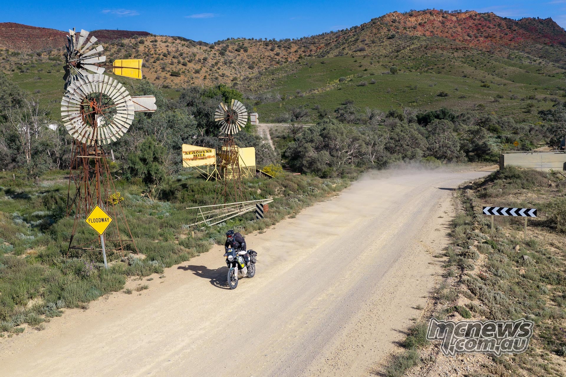 Husqvarna Norden rider passes two windmills beside a gravel road, with green hills and rocky ridges behind.