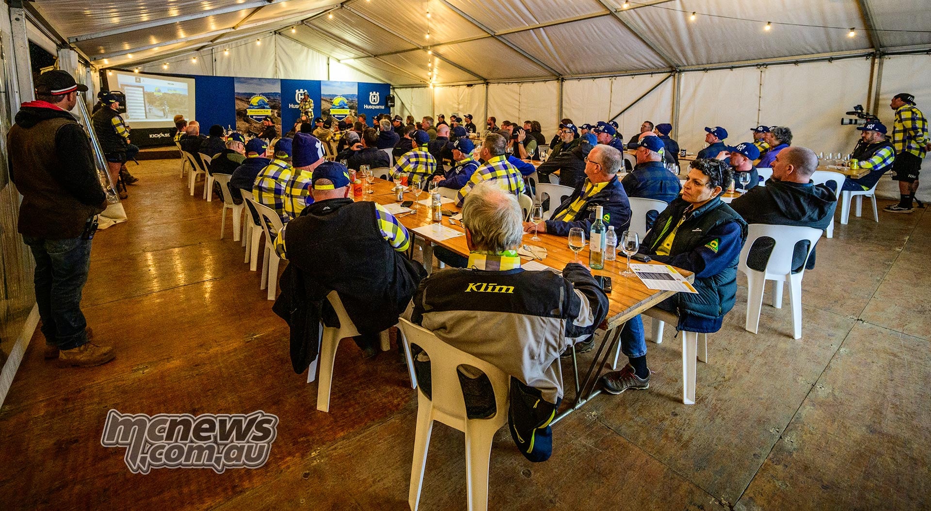 Husqvarna Trek participants sit at long tables inside a marquee while a speaker stands beside a presentation screen.