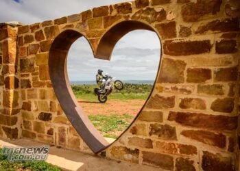 Husqvarna 701 Enduro rider performs a wheelie, viewed through a heart-shaped opening in a stone wall.
