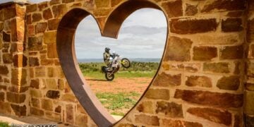 Husqvarna 701 Enduro rider performs a wheelie, viewed through a heart-shaped opening in a stone wall.