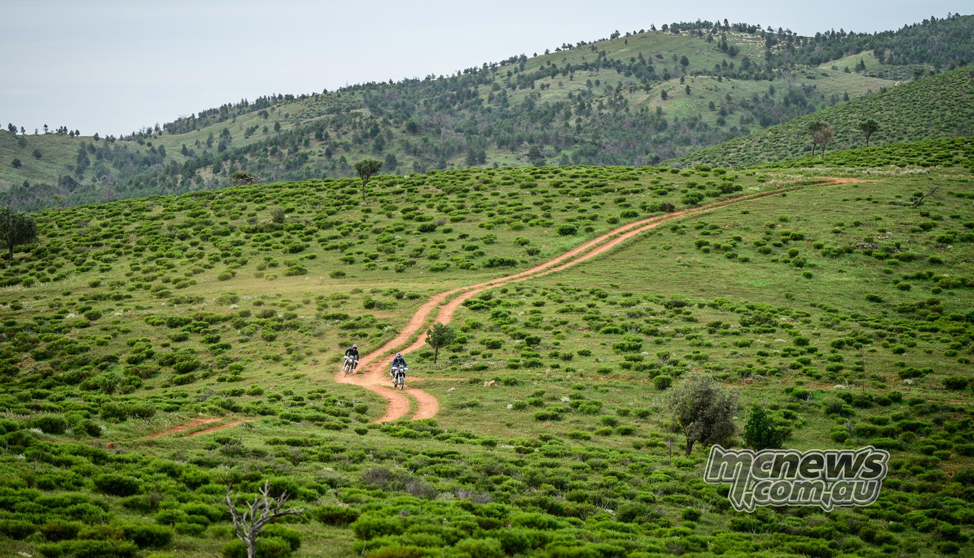 Two adventure riders follow a red dirt track through vivid green scrub and rolling hills.