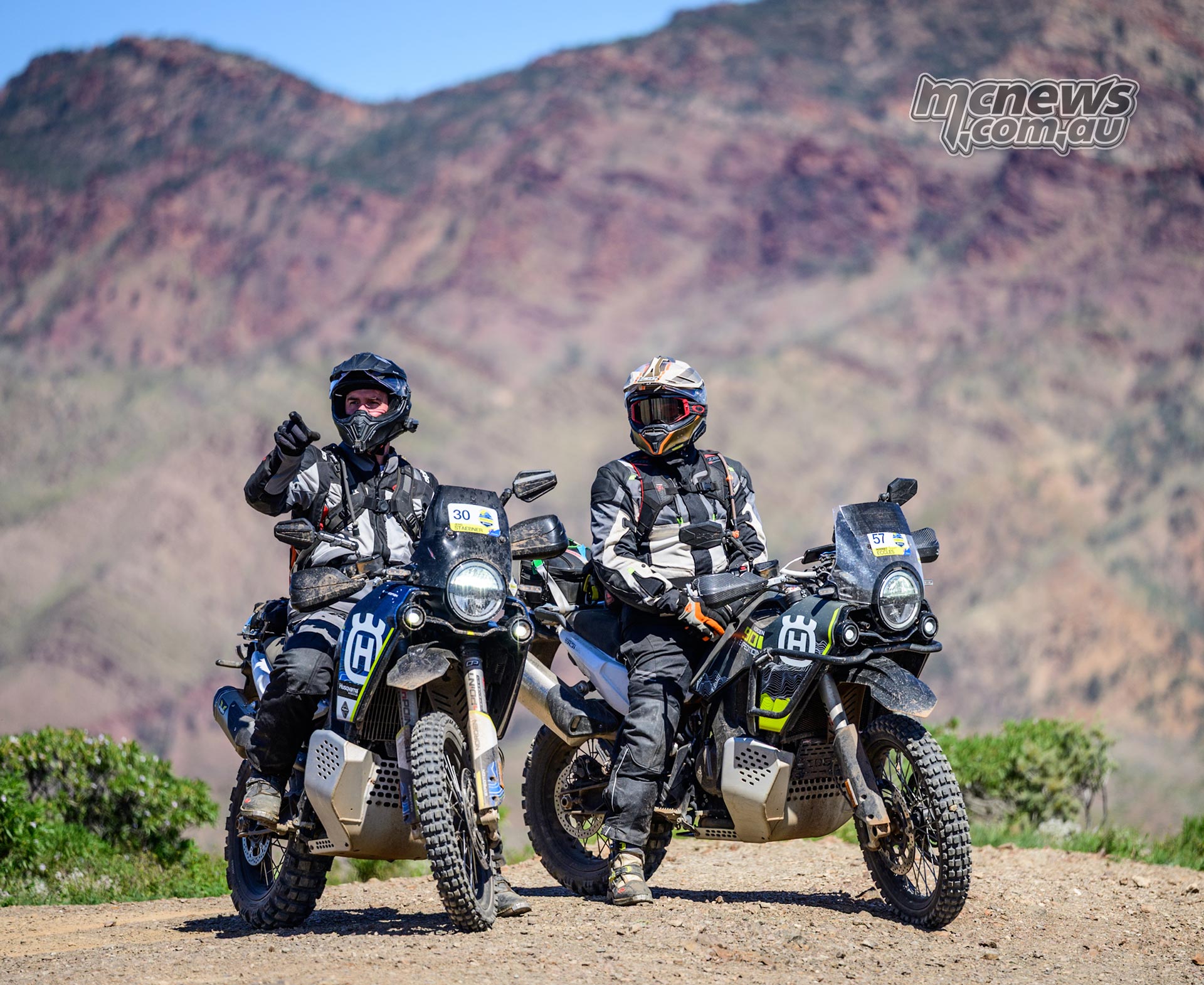 Two Husqvarna Norden 901 riders stopped beside each other on a dirt track with rocky mountains behind.
