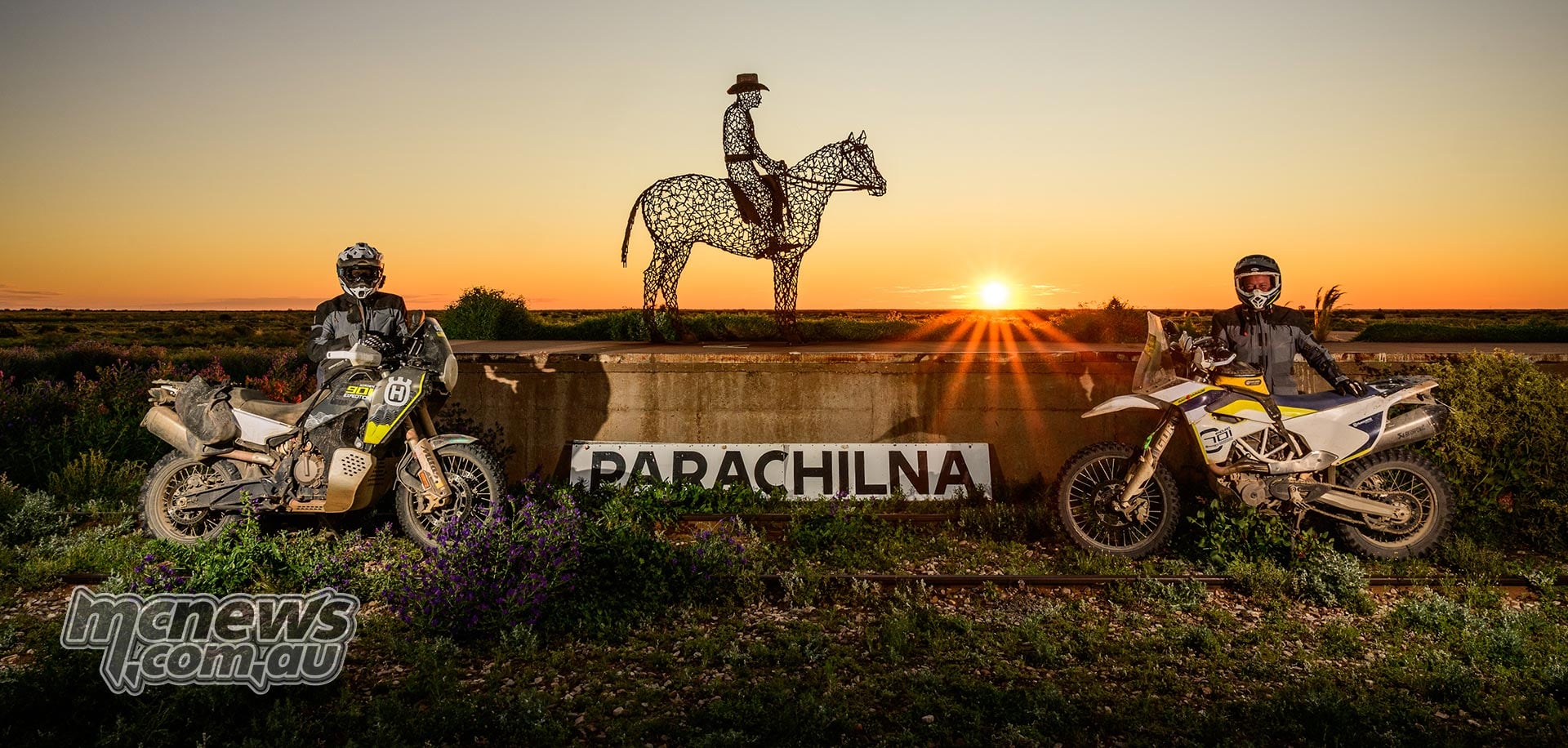 Two Husqvarna riders beside the Parachilna sign and a horse-and-rider sculpture at sunset.