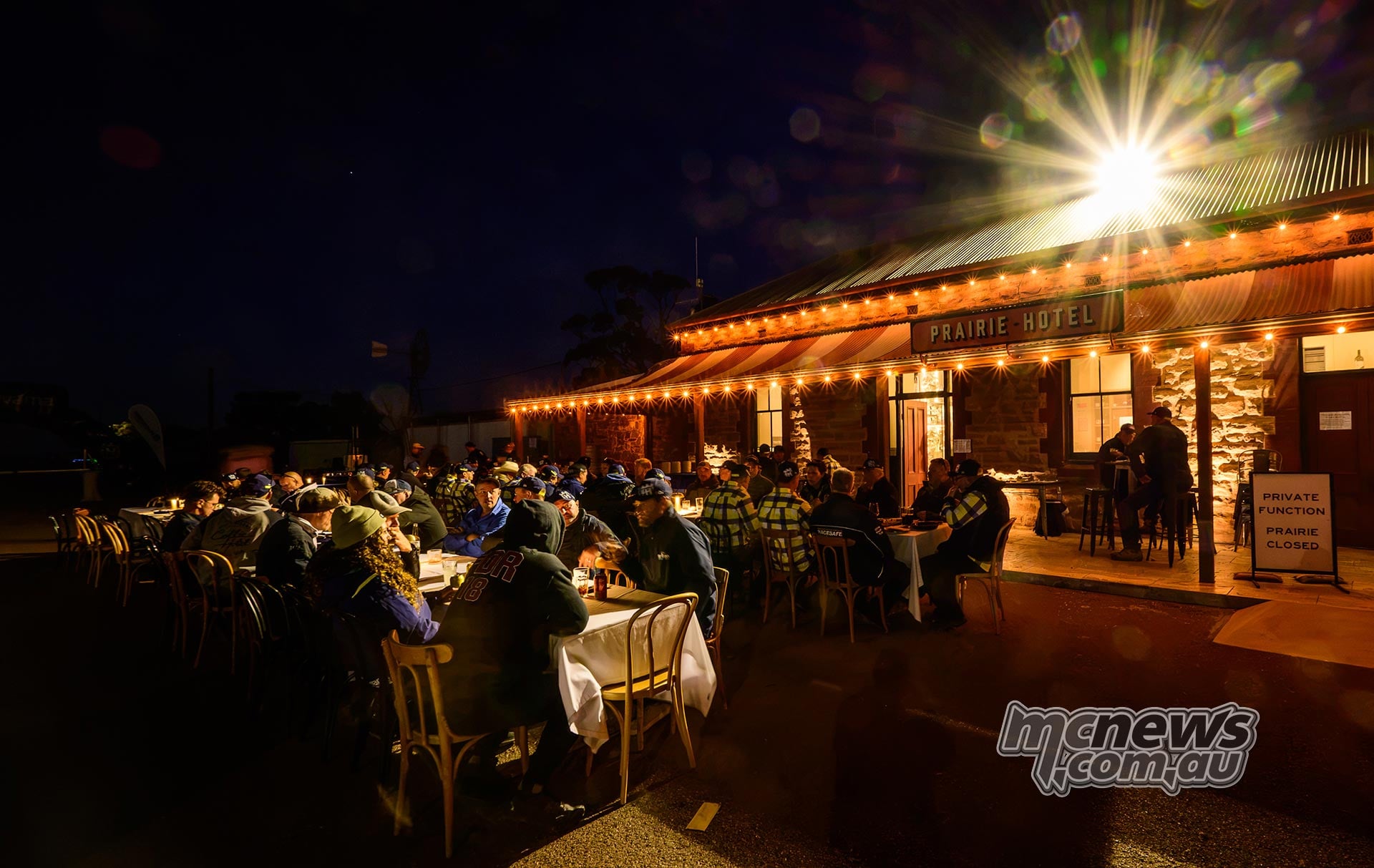 Husqvarna Trek participants dine outdoors at long tables beneath lights outside the Prairie Hotel at night.