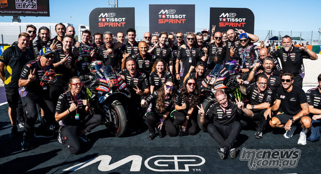 Aprilia riders and team members celebrate together around their motorcycles in Misano parc ferme.