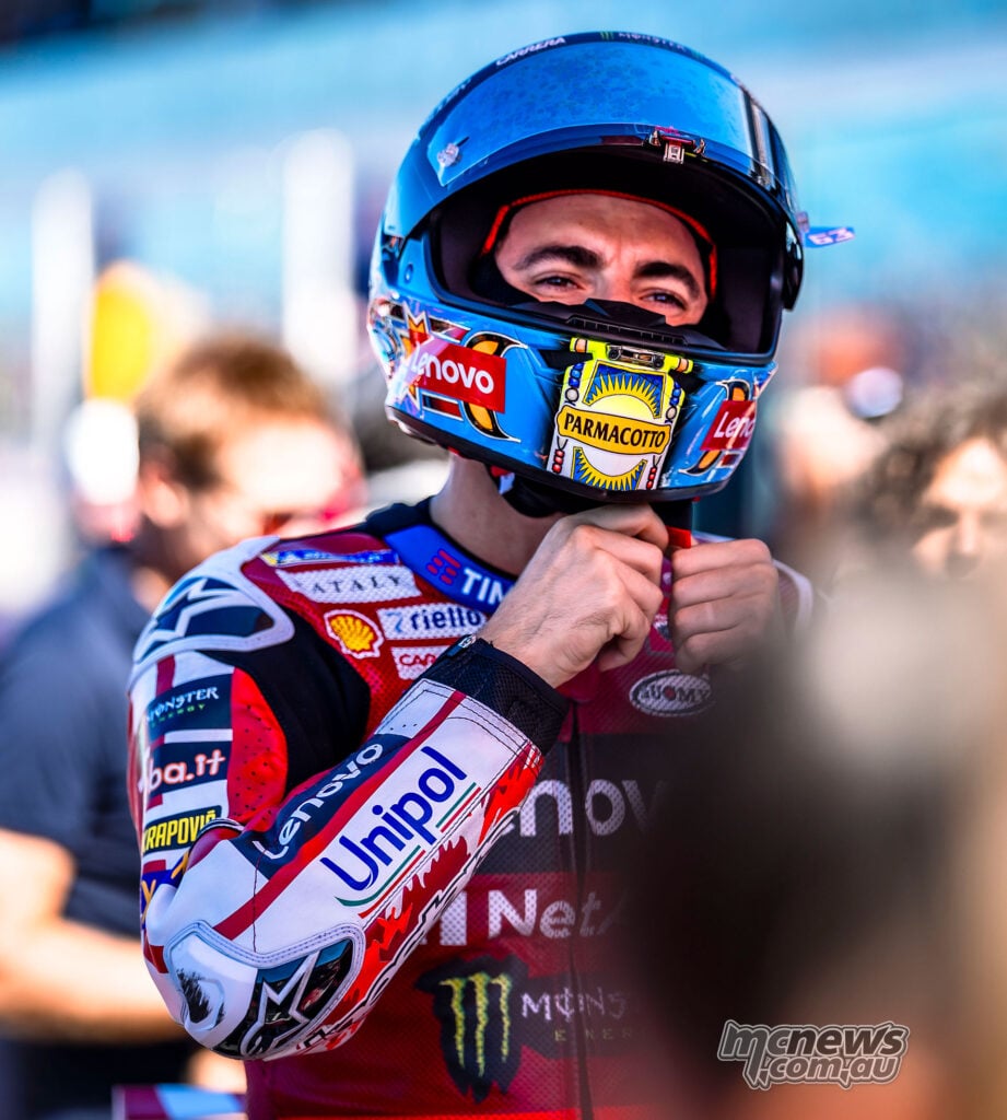 Francesco Bagnaia holds the strap beneath his blue helmet, with the visor raised and his Ducati Lenovo leathers visible.