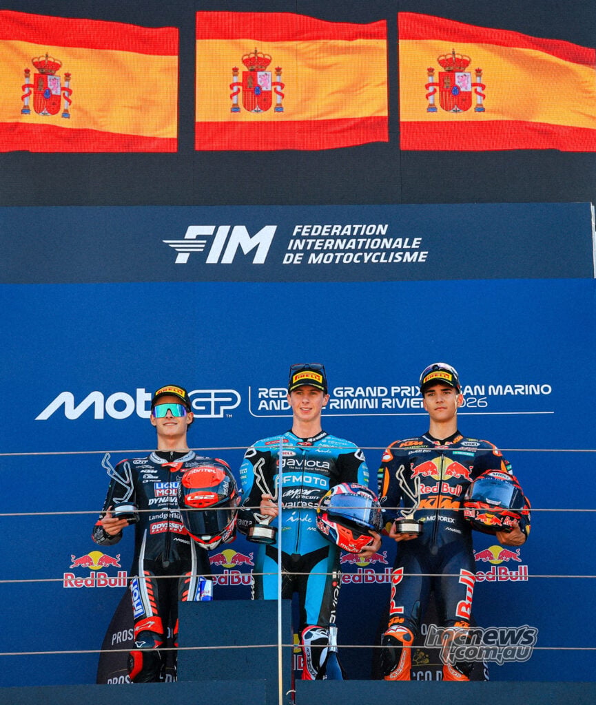 David Almansa, Maximo Quiles and Brian Uriarte hold trophies and helmets beneath three Spanish flags on the Misano podium.