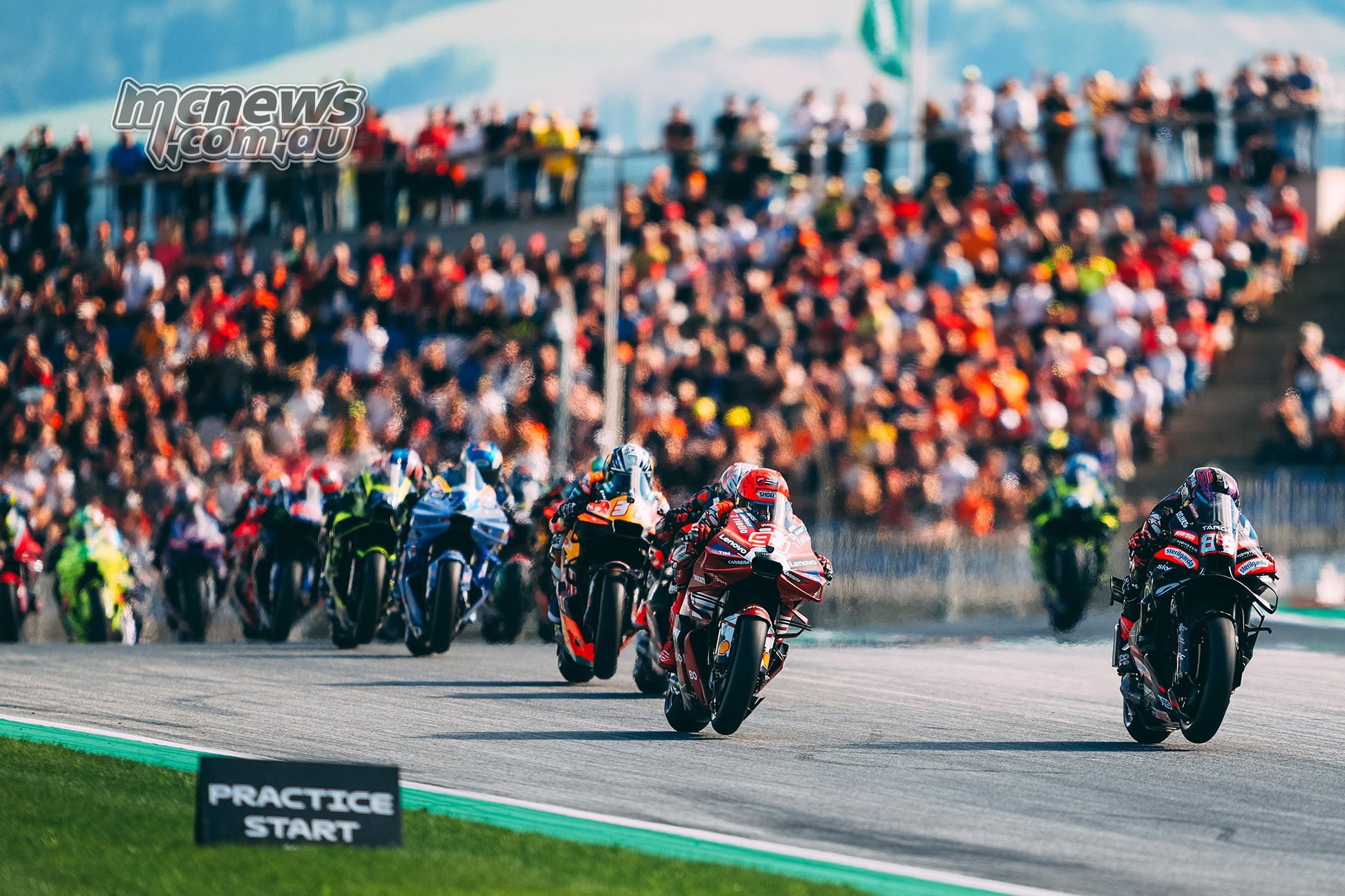 MotoGP riders accelerate away at the start of the Austrian Sprint, with spectators behind them.