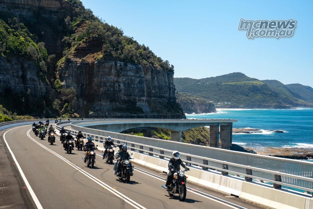 Group of Royal Enfield riders crossing Sea Cliff Bridge, with coastal cliffs to the left and the ocean to the right.