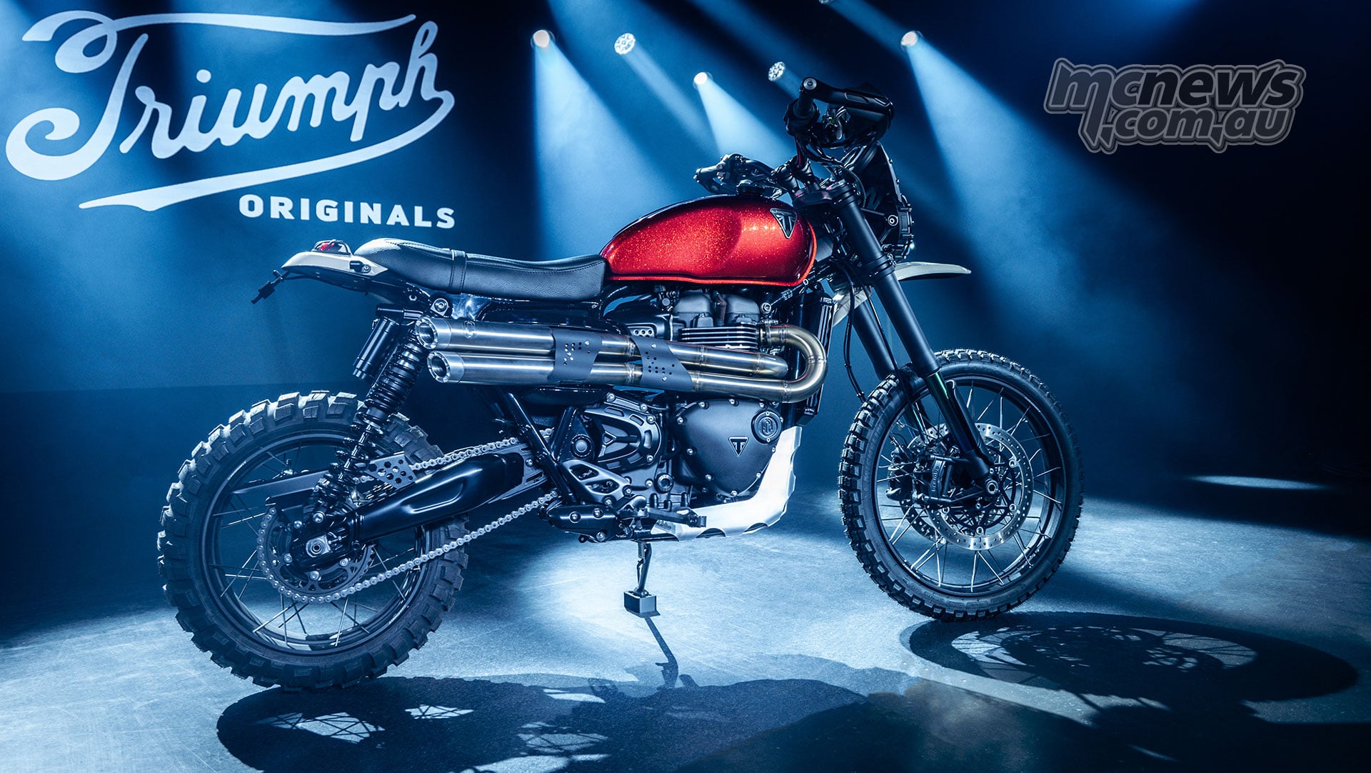 FCR Original Red Dust Triumph Scrambler 1200 XE with a red tank and high twin exhausts, displayed under blue spotlights.