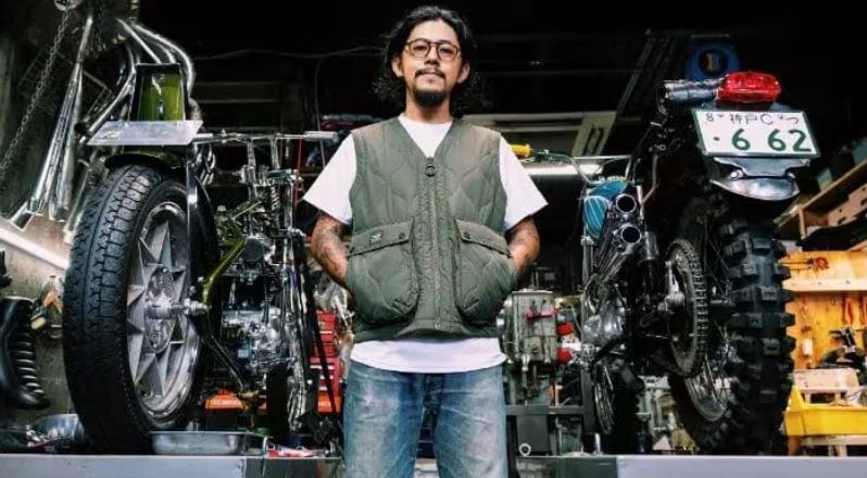 Satoshi of Fonk Motorcycles standing between custom motorcycles in his workshop, wearing a green vest.